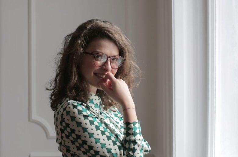 Brunetka w okularach z czarnymi oprawkami ubrana w koszulę w biało-zieloną kratkę stoi bokiem i trzyma rękę na twarzy nieśmiało patrząc