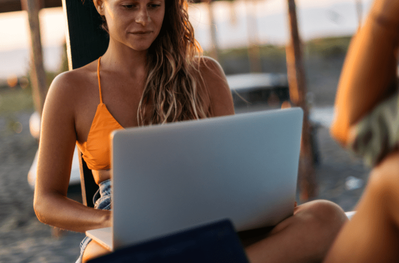 Cyfrowi nomadzi, digital nomads | kobieta siedzi na plaży i pracuje przy komputerze