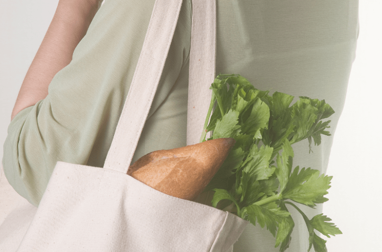 Ekologia i oszczędzanie/ Kobieta z płócienną torbą na ramieniu. W torbie są zakupy - wystają z niej bagietka i zielona nać.