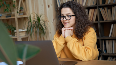Kobieta w żółtym swetrze siedzi przy stole i patrzy na ekran laptopa.
