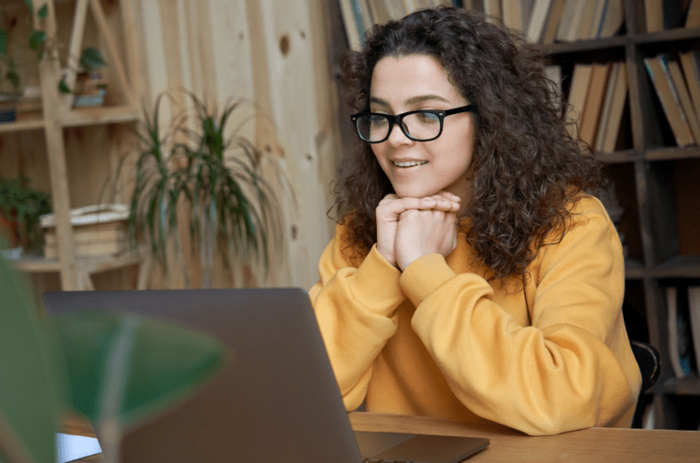 Kobieta w żółtym swetrze siedzi przy stole i patrzy na ekran laptopa.