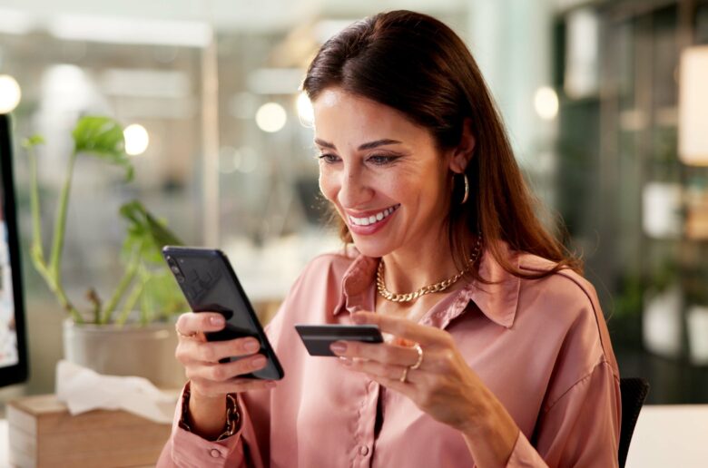 Kobieta trzymająca w jednej dłoni kartę kredytową, a w drugiej telefon komórkowy.