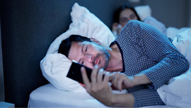 Mężczyzna leżący w łóżku w ciemnym pokoju, spoglądający na telefon komórkowy. Obok niego, w tle, widać osobę śpiącą pod kołdrą. Mężczyzna wygląda na zamyślonego lub skupionego, trzymając telefon blisko twarzy. Scena sugeruje tajemniczą lub intymną atmosferę.