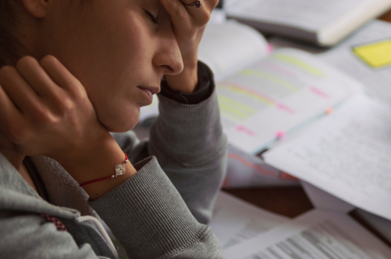 Zmęczona i smutna kobieta, trzyma się za czoło. Pracuje przy biurku pełnym dokumentów.